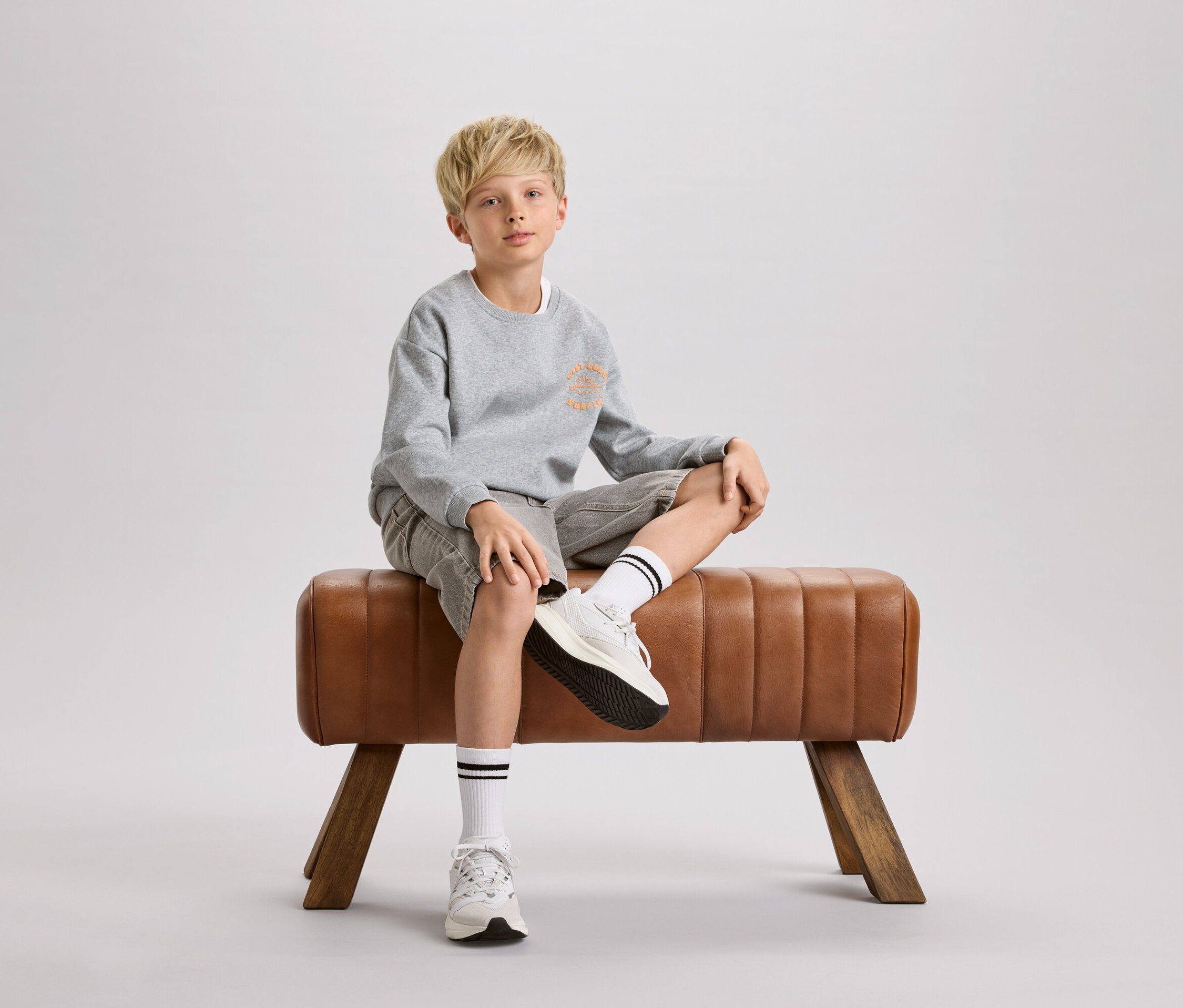 Un garçon blond est assis sur un cheval d'arçons marron. Il porte un sweat-shirt gris, un short et des chaussettes blanches à rayures noires.