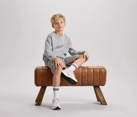 Un garçon blond est assis sur un cheval d'arçons marron. Il porte un sweat-shirt gris, un short et des chaussettes blanches à rayures noires.