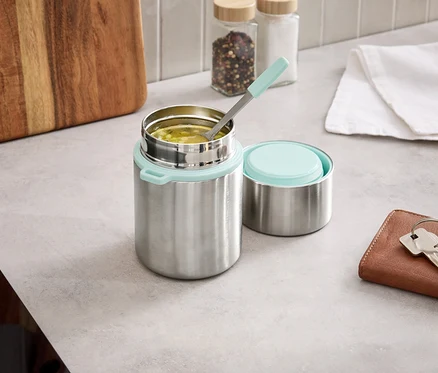Un thermos argenté avec un couvercle bleu clair est ouvert sur un comptoir de cuisine, à côté du couvercle se trouve une cuillère dans la nourriture.