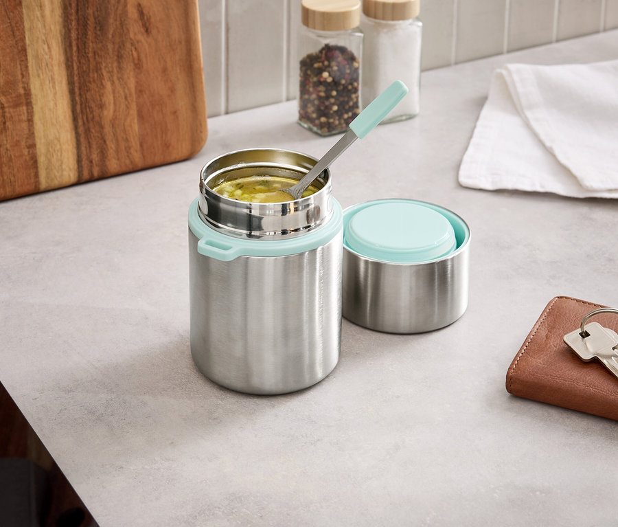 Un thermos argenté avec un couvercle bleu clair est ouvert sur un comptoir de cuisine, à côté du couvercle se trouve une cuillère dans la nourriture.