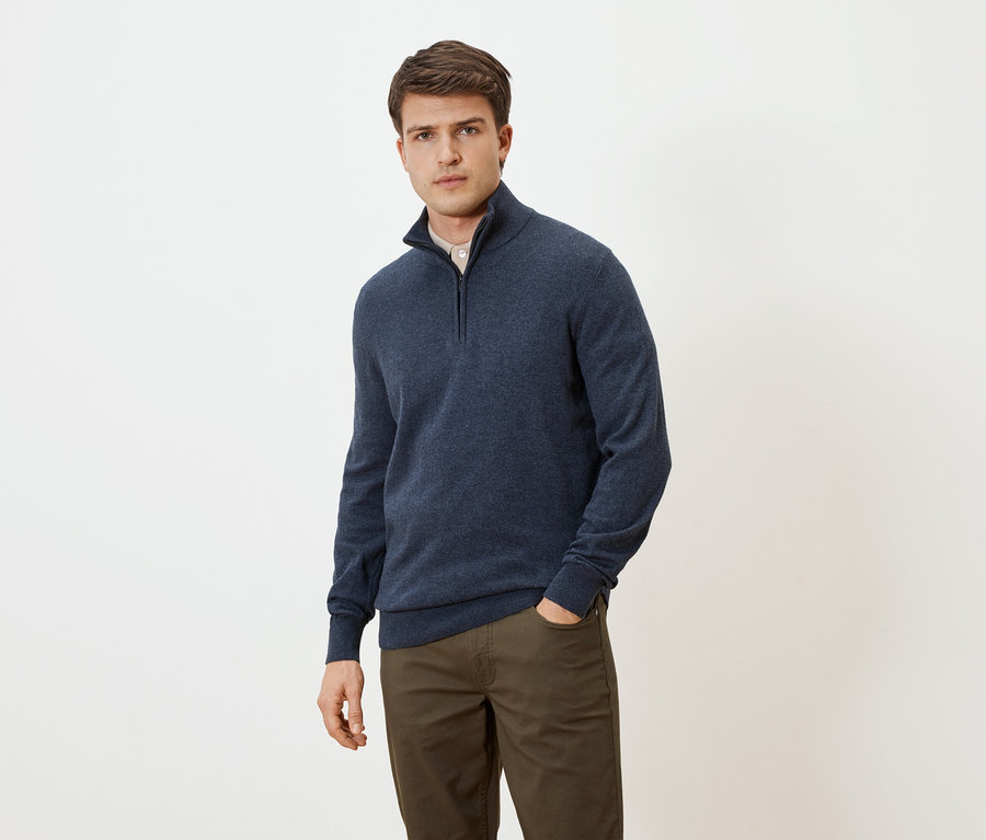 Homme portant un pull bleu zippé et un pantalon marron, une main dans la poche, devant un fond neutre.