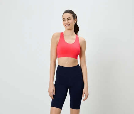 Une femme souriante portant un soutien-gorge de sport rose et un short de cycliste bleu marine sur fond blanc.