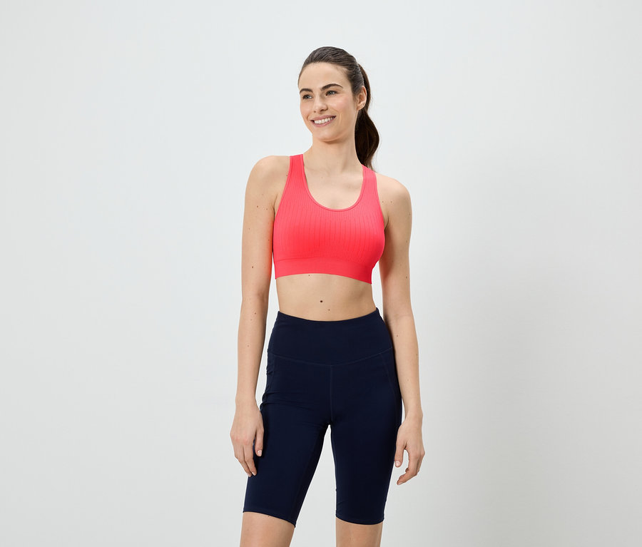 Une femme souriante portant un soutien-gorge de sport rose et un short de cycliste bleu marine sur fond blanc.