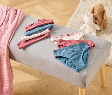Sur un banc gris reposent des culottes pliées et dépliées dans les tons rose, bleu et blanc, à côté d'une couverture rose et de peluches.