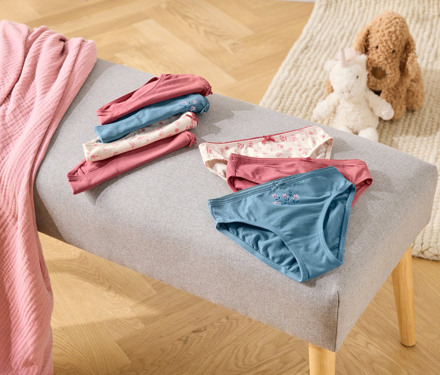 Sur un banc gris reposent des culottes pliées et dépliées dans les tons rose, bleu et blanc, à côté d'une couverture rose et de peluches.