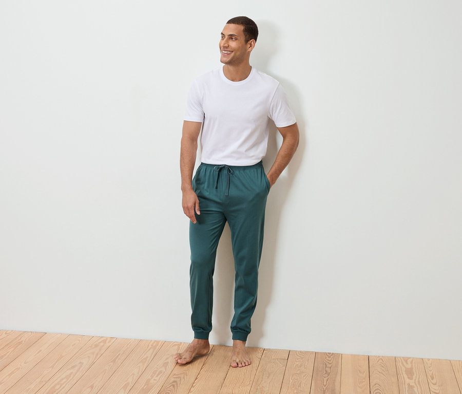 Un homme se tient pieds nus, la main dans la poche, devant un mur blanc. Il porte un t-shirt blanc et un pantalon de jogging bleu pétrole.