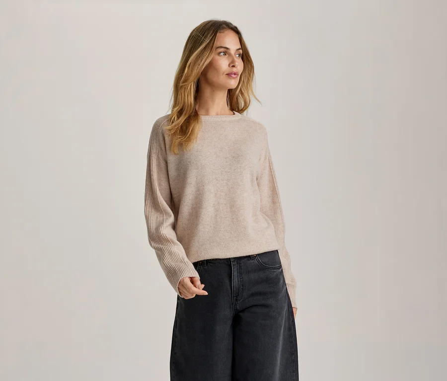 Une femme aux longs cheveux blonds ondulés se tient devant un fond clair, regardant vers sa droite. Elle porte un pull ras-du-cou beige clair à manches côtelées et un jean gris foncé à jambes larges.
