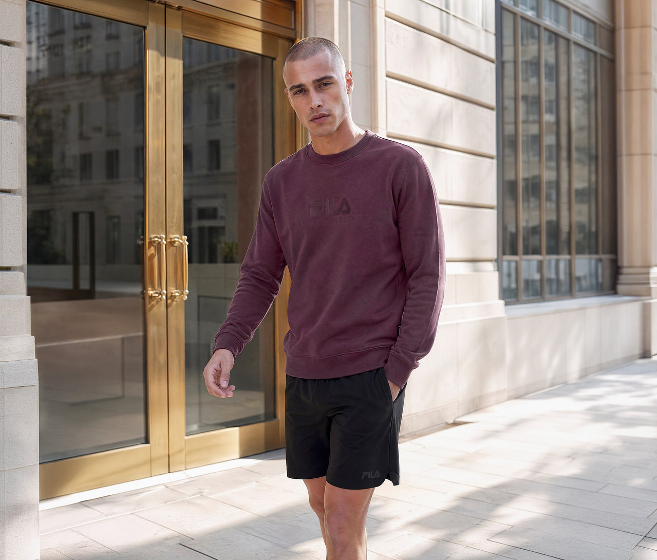 Homme en sweat-shirt Fila bordeaux et short Fila noir debout devant un bâtiment moderne avec des portes vitrées.
