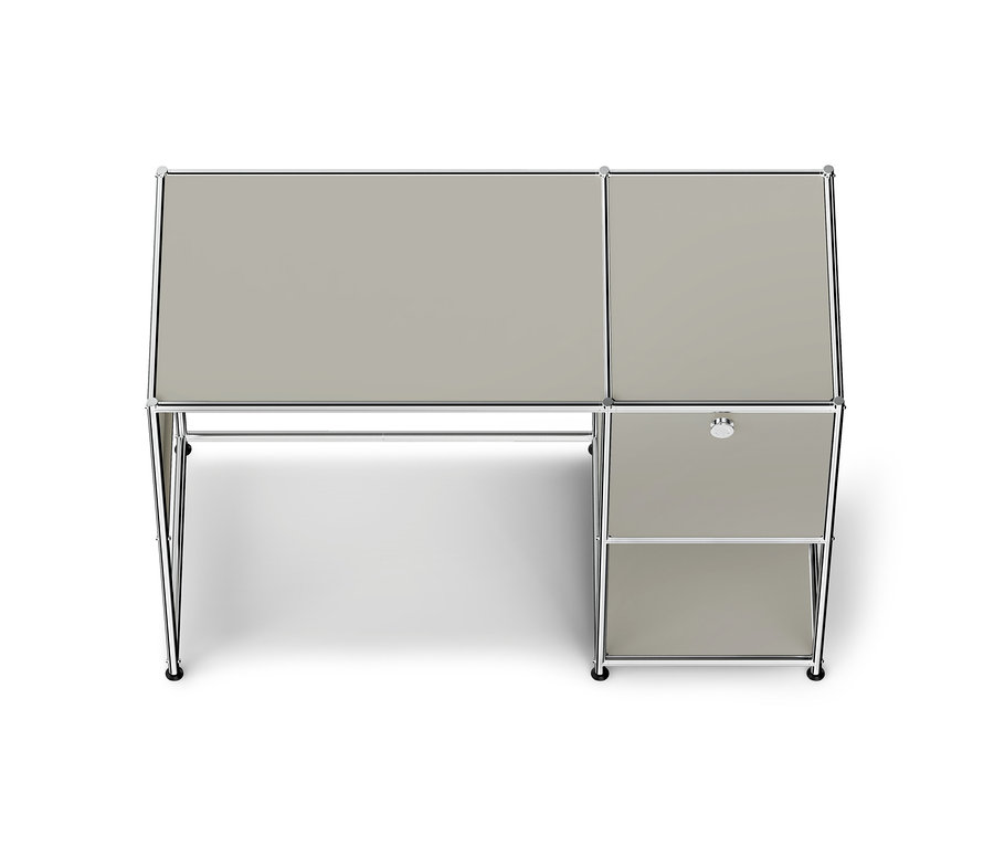 Un bureau modulaire moderne gris clair avec un cadre chromé, vu de dessus. Le bureau a un grand plateau principal à gauche et un plus petit plateau à droite, qui est placé au-dessus d'une unité avec un tiroir unique et une étagère ouverte. Le bureau est soutenu par des pieds chromés avec des roulettes.