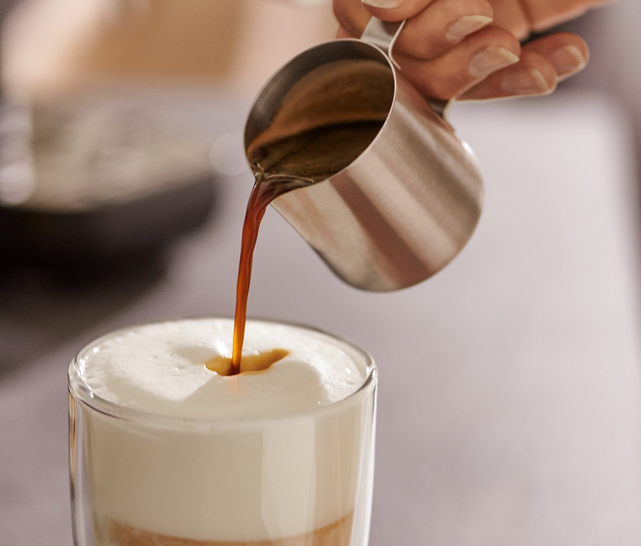 Une femme verse de l'expresso d'un pichet à expresso en inox (80 ml) dans un verre de lait.