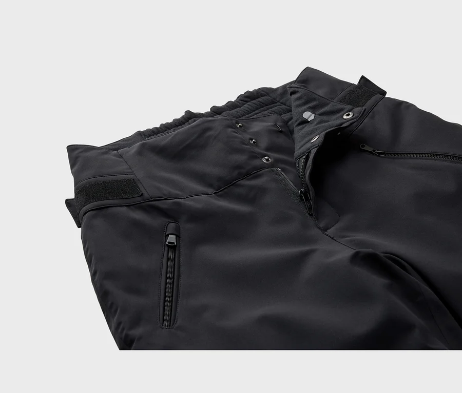 Gros plan sur la partie supérieure d'un pantalon technique noir, montrant une braguette partiellement ouverte avec des boutons-pression, une taille élastique, une poche zippée et une sangle Velcro sur le côté.