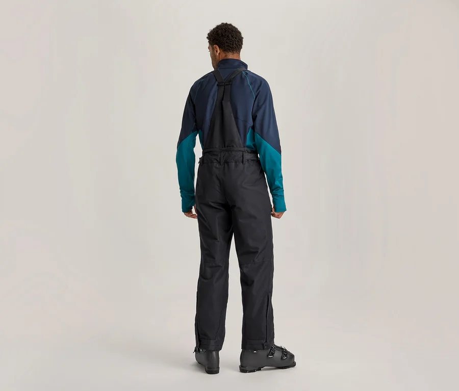 Homme de dos en tenue de ski, portant un haut à manches longues bleu foncé et turquoise, une salopette de ski noire et des chaussures de ski noires.
