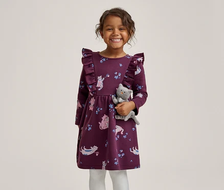 Une petite fille souriante vêtue d'une robe violette à volants avec un motif de chats, tenant un chat en peluche gris.