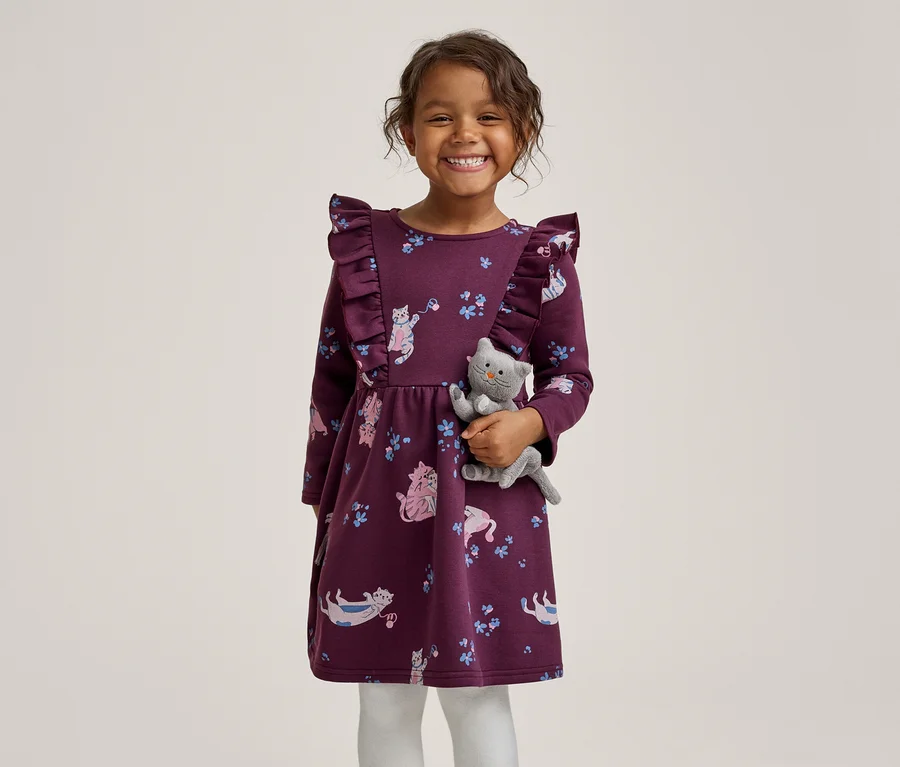 Une petite fille souriante vêtue d'une robe violette à volants avec un motif de chats, tenant un chat en peluche gris.