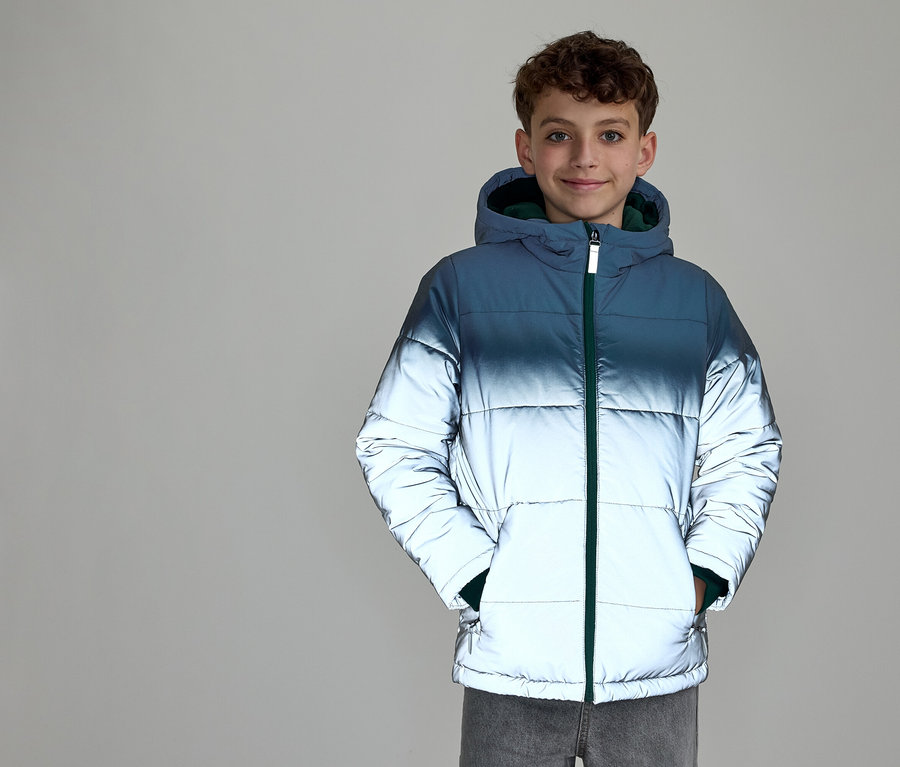Un jeune garçon souriant aux cheveux bouclés porte une doudoune à capuche. La veste présente un dégradé de couleur, passant du bleu foncé en haut à l'argent réfléchissant en bas, avec une fermeture éclair verte. Ses mains sont dans les poches de la veste.