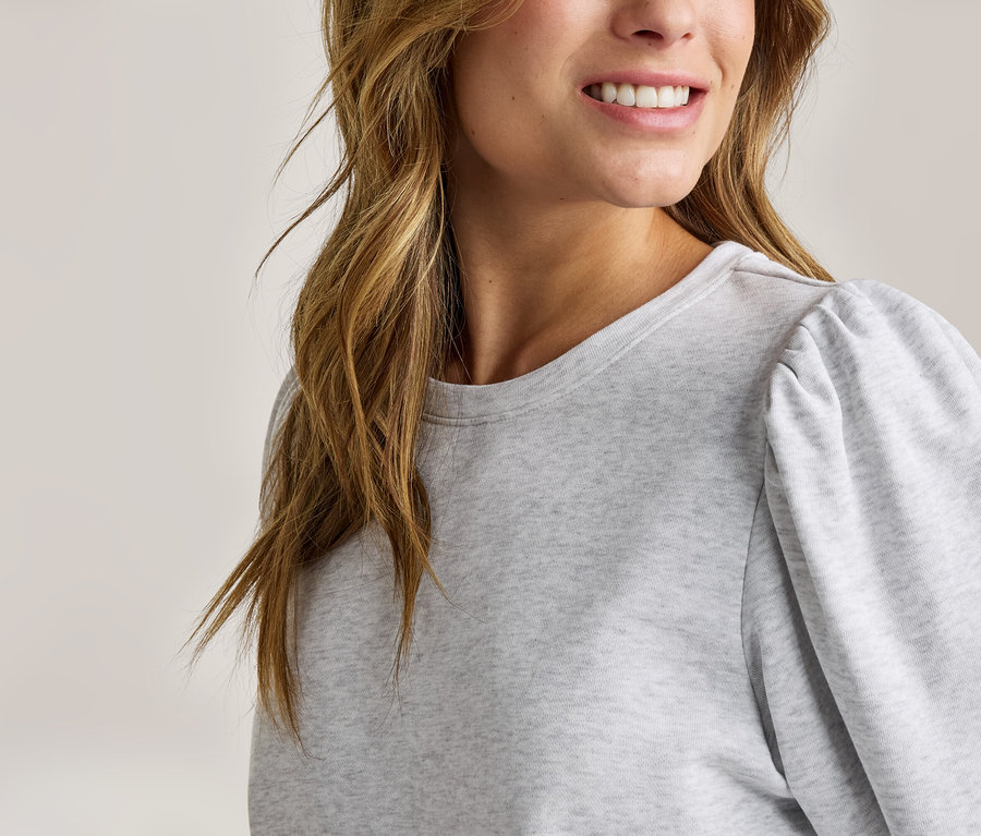 Gros plan d'une femme souriante aux longs cheveux bruns ondulés, portant un sweat-shirt gris clair à manches bouffantes.