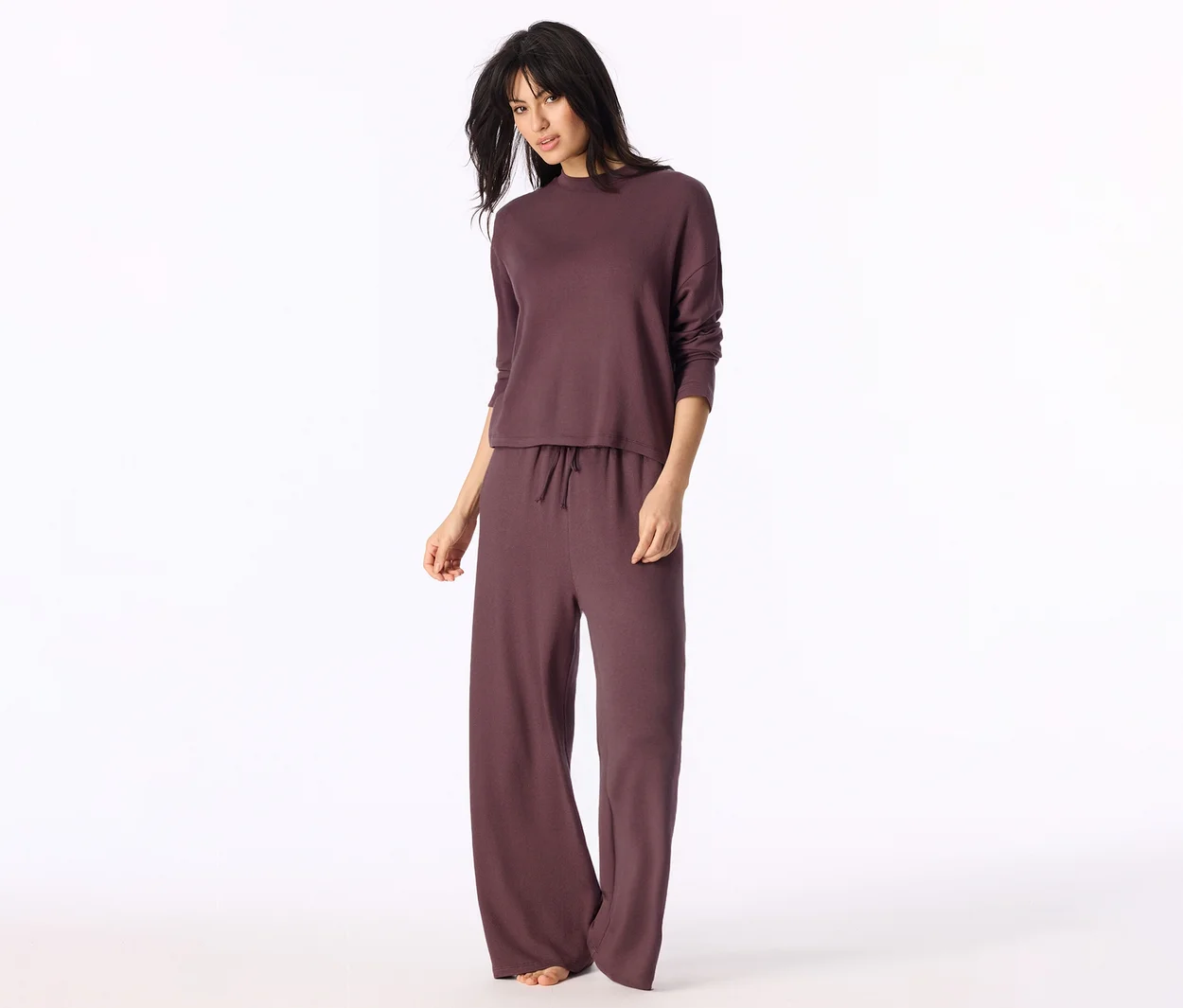 Femme en ensemble loungewear prune foncée: haut à manches longues et pantalon large à cordon de serrage. Elle est pieds nus et regarde l'appareil photo.