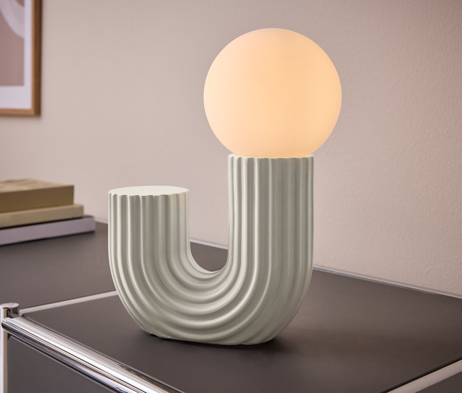 Une lampe de table en forme de U avec une base nervurée et une ampoule sphérique lumineuse, posée sur un meuble sombre. Des livres et un tableau sont en arrière-plan.