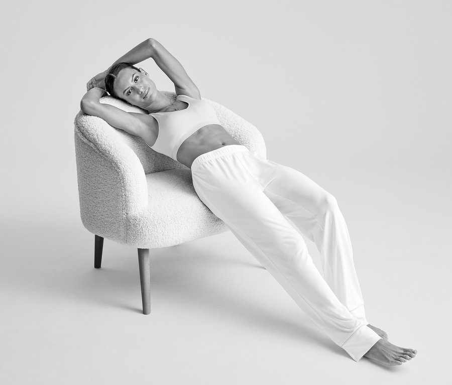 Une femme vêtue d'une brassière sans coutures crème et d'un pantalon clair est allongée sur un fauteuil texturé, les bras derrière la tête.
