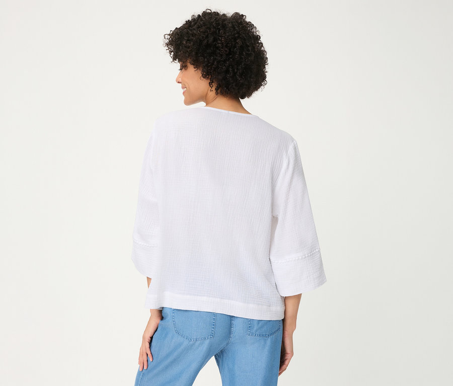 Femme de dos, légèrement tournée, portant une blouse tissée Olsen blanche texturée à manches 3/4 et un pantalon bleu clair.