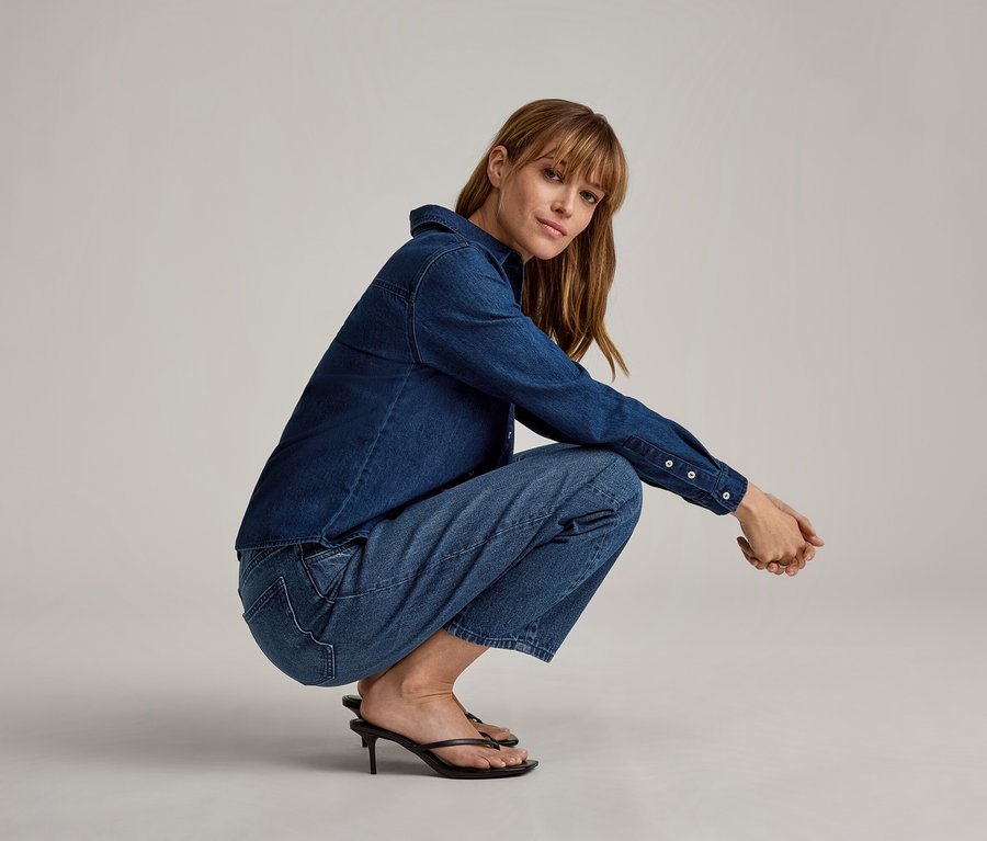Femme accroupie, portant une chemise en jean bleu foncé, un jean bleu clair et des sandales à talons aiguilles noires.