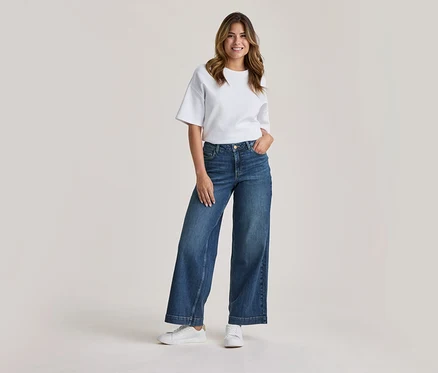Femme souriante debout, portant un t-shirt blanc, un jean bleu large et des baskets blanches, une main dans la poche.