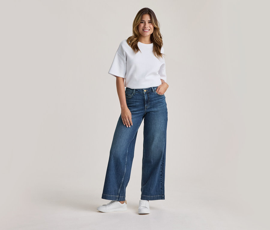 Femme souriante debout, portant un t-shirt blanc, un jean bleu large et des baskets blanches, une main dans la poche.