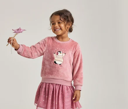Fille souriante portant un pull rose duveteux avec un pingouin et une jupe rose à étoiles, tenant une baguette étoile rose.