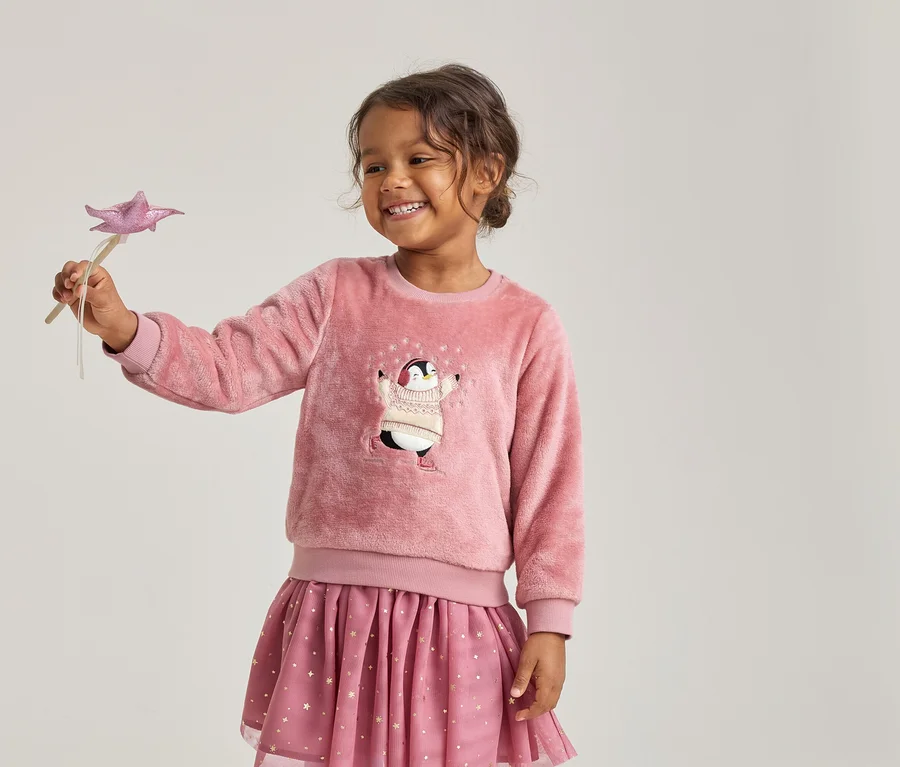Fille souriante portant un pull rose duveteux avec un pingouin et une jupe rose à étoiles, tenant une baguette étoile rose.
