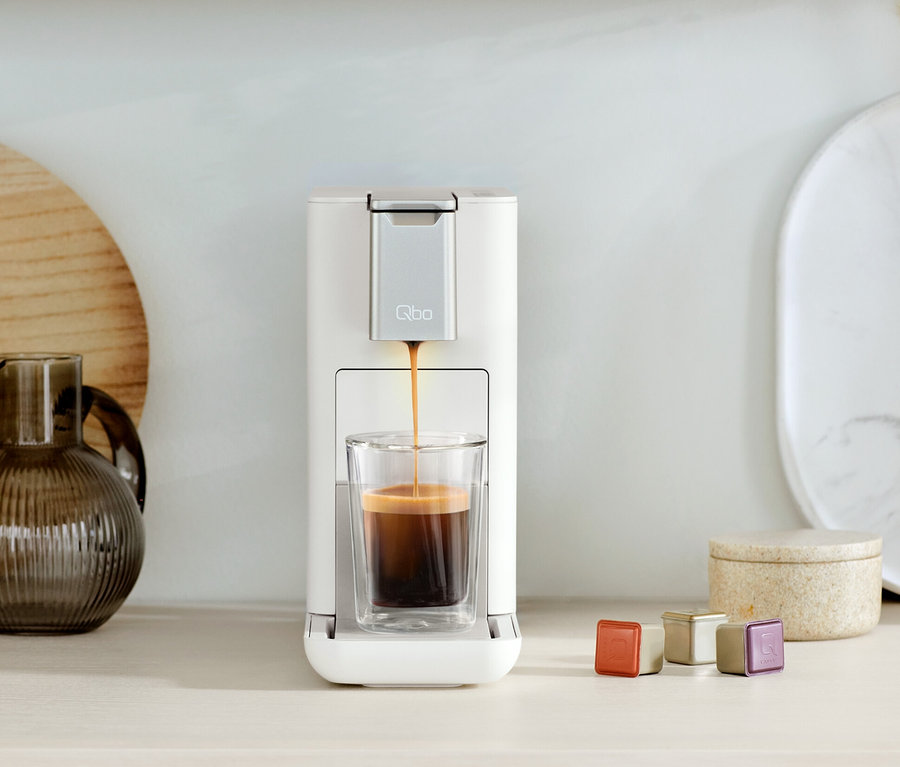 La machine à café Qbo ICONIC Dark Chrome et Qbo ESSENTIAL Bright Sand remplit un verre de café. Des capsules sont à côté.