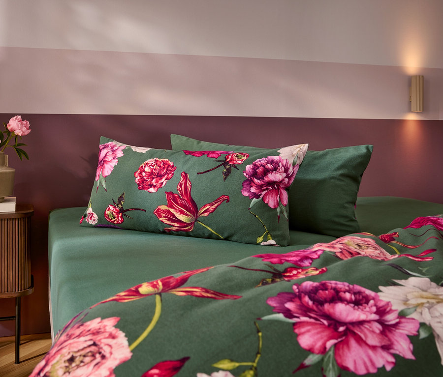 Un lit avec une parure de lit vert foncé à motifs de grandes fleurs roses et rouges, et un oreiller vert uni. À gauche, une table de chevet avec un vase rempli de pivoines roses. Le mur est bicolore, rose et rouge foncé.