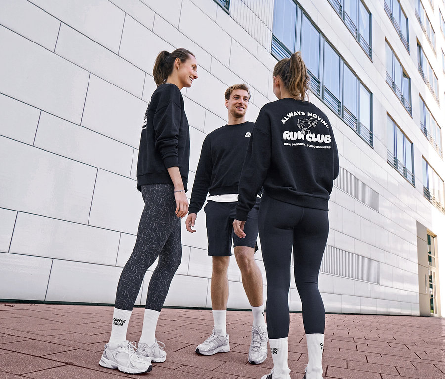 Trois personnes en sweatshirts noirs et tenues de sport discutent. Une femme porte des leggings de sport « ActivePower » gris-noir et des chaussettes de tennis Unisex Coffee Crew. Un homme porte un short de sport noir et des chaussettes blanches. Une autre femme porte des leggings de sport « ActivePower » noirs et un sweatshirt avec le logo « RUN CLUB ».
