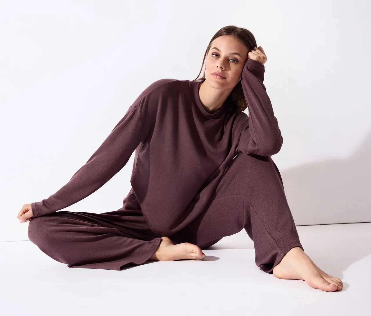 Femme assise sur un sol blanc, vêtue d'un haut à manches longues et d'un pantalon large assorti de couleur prune foncé, appuyée sur une main et regardant l'appareil photo.