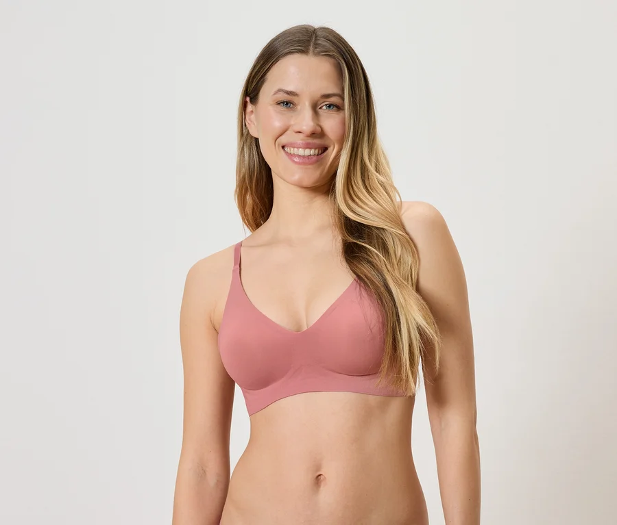 Femme souriante aux longs cheveux blonds portant un soutien-gorge rose sur fond clair.