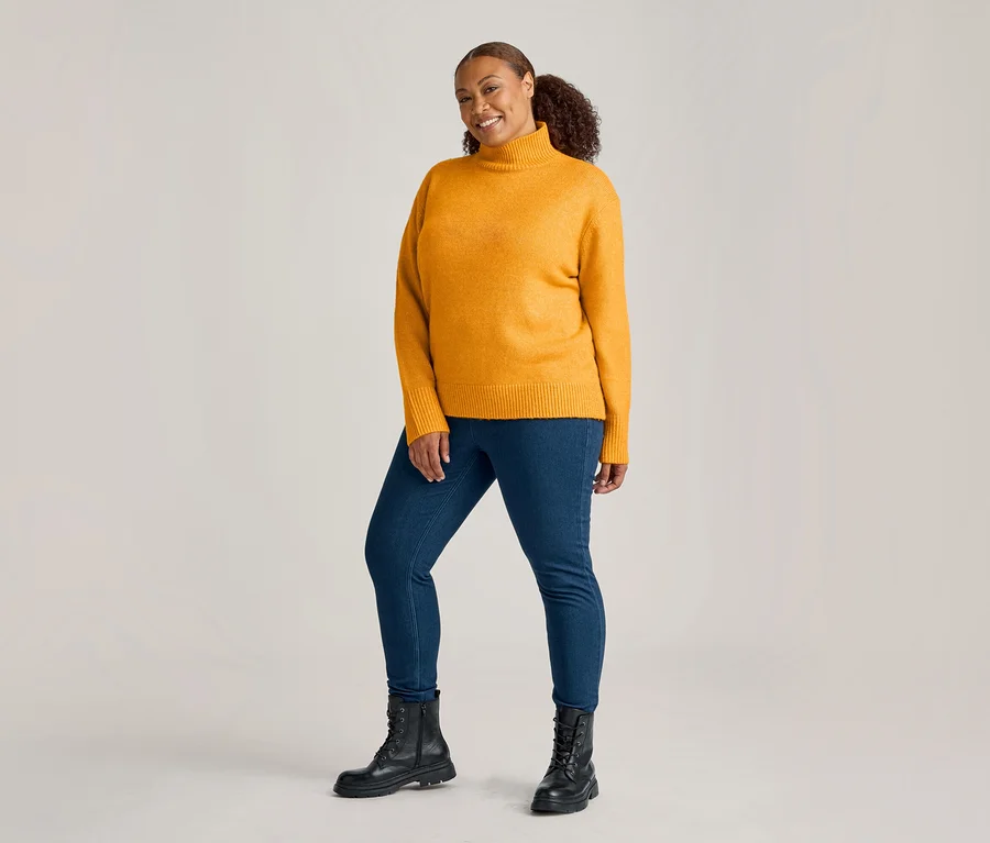 Femme souriante en pull col roulé jaune moutarde, jeggings denim bleu moyen et bottes lacées noires, regardant la caméra.