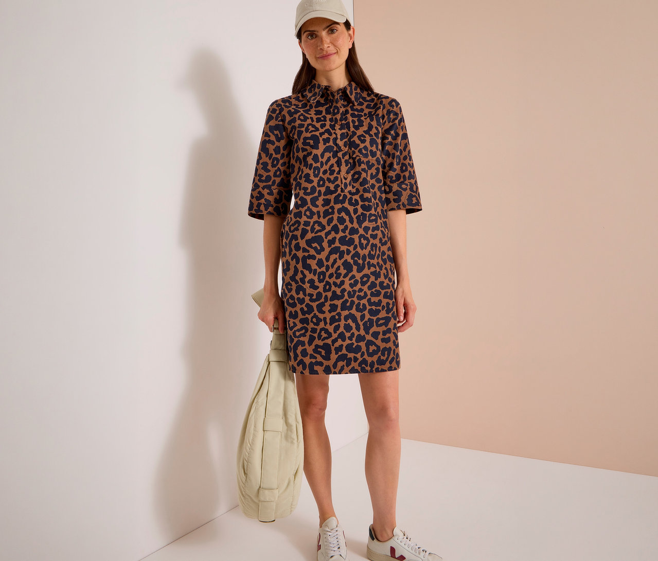 Femme portant une robe en velours côtelé CECIL à imprimé léopard marron et noir, une casquette de baseball beige et des baskets blanches, tenant un sac matelassé vert clair.
