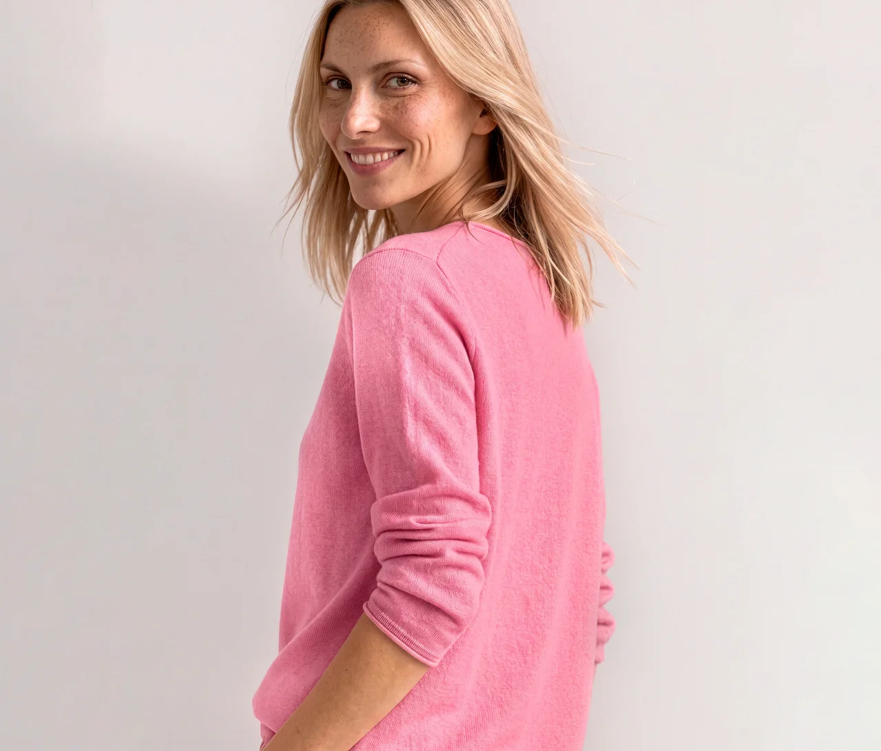 Une femme souriante aux cheveux blonds et aux taches de rousseur regarde par-dessus son épaule, vêtue d'un pull en maille Street One rose pâle.