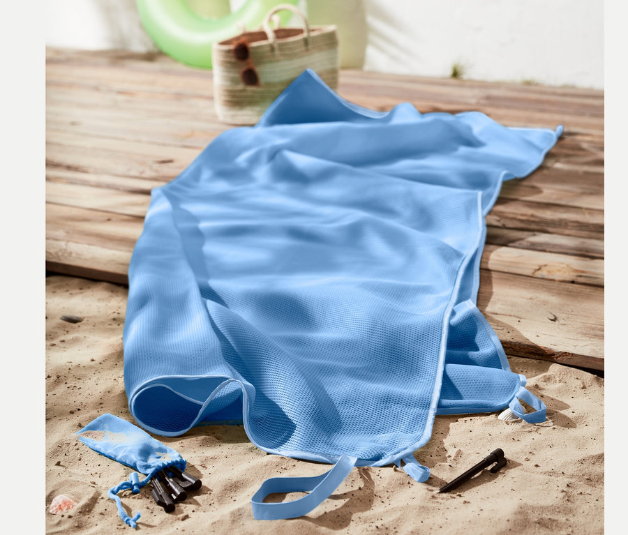 Une couverture de plage anti-sable bleu clair est posée sur le sable à côté d'un sac de plage et d'une bouée.