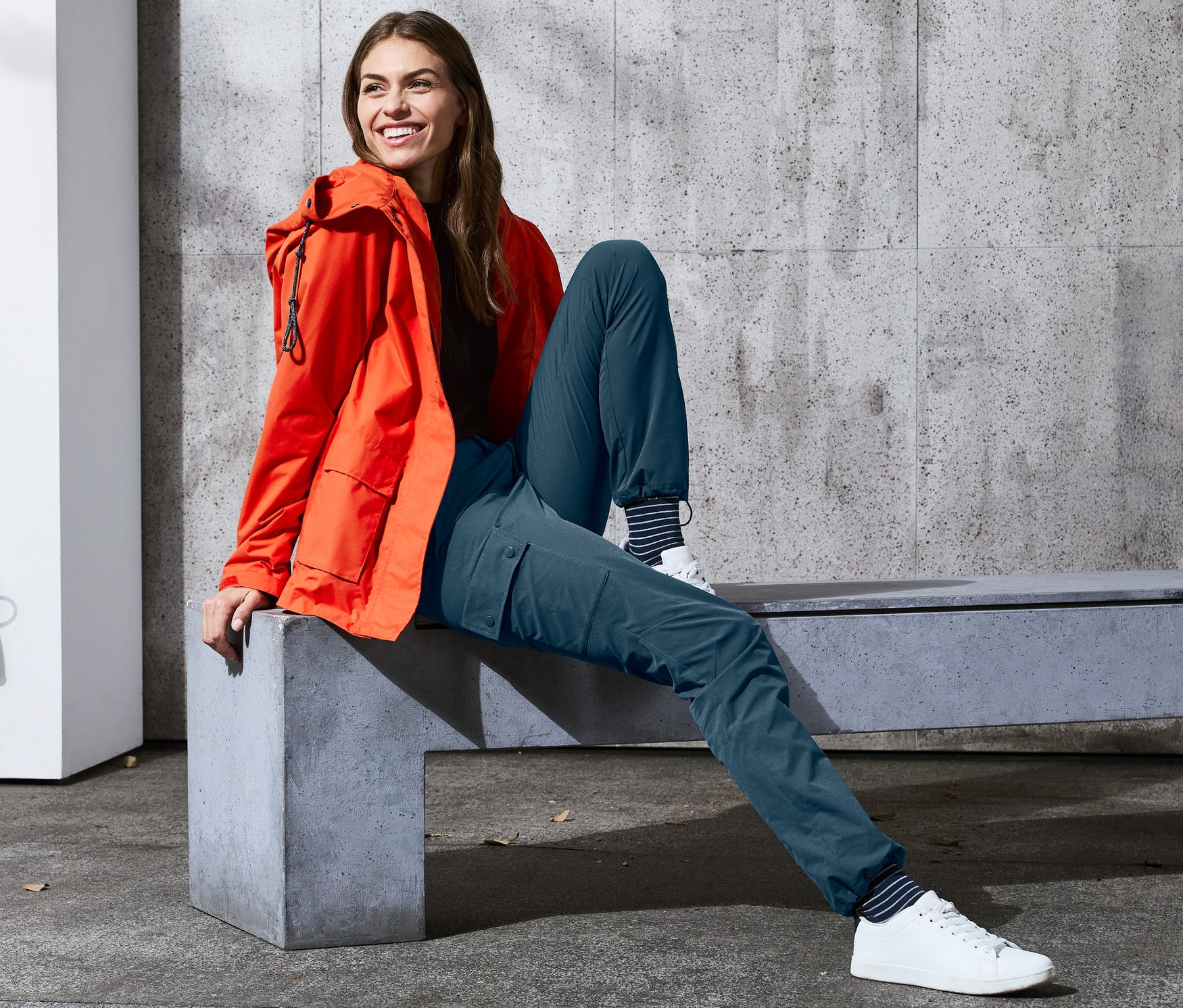 Une femme assise sur un banc porte un pantalon softshell et une veste orange.