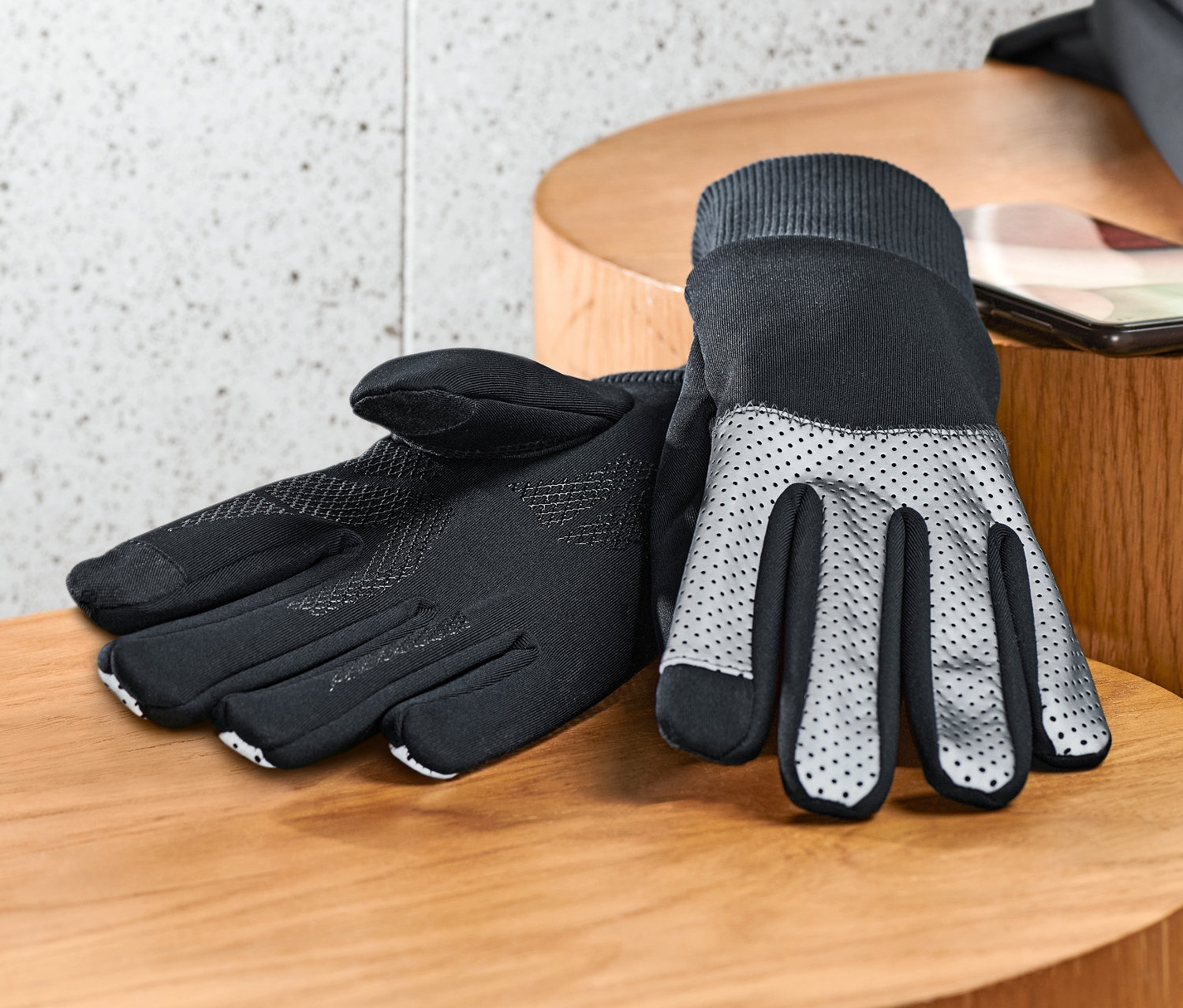 Des gants noirs coupe-vent avec des bordures réfléchissantes sont posés sur une table en bois à côté d'un téléphone portable et d'une veste noire.