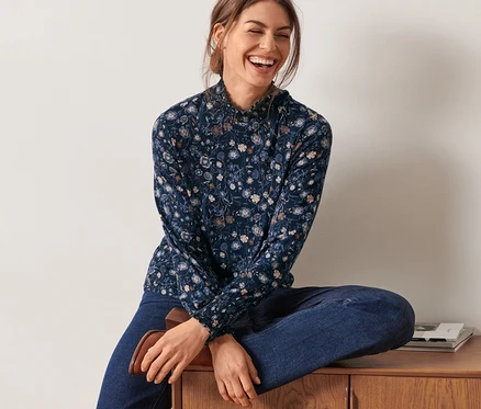Femme assise sur une commode en bois portant une blouse à fleurs avec col montant.