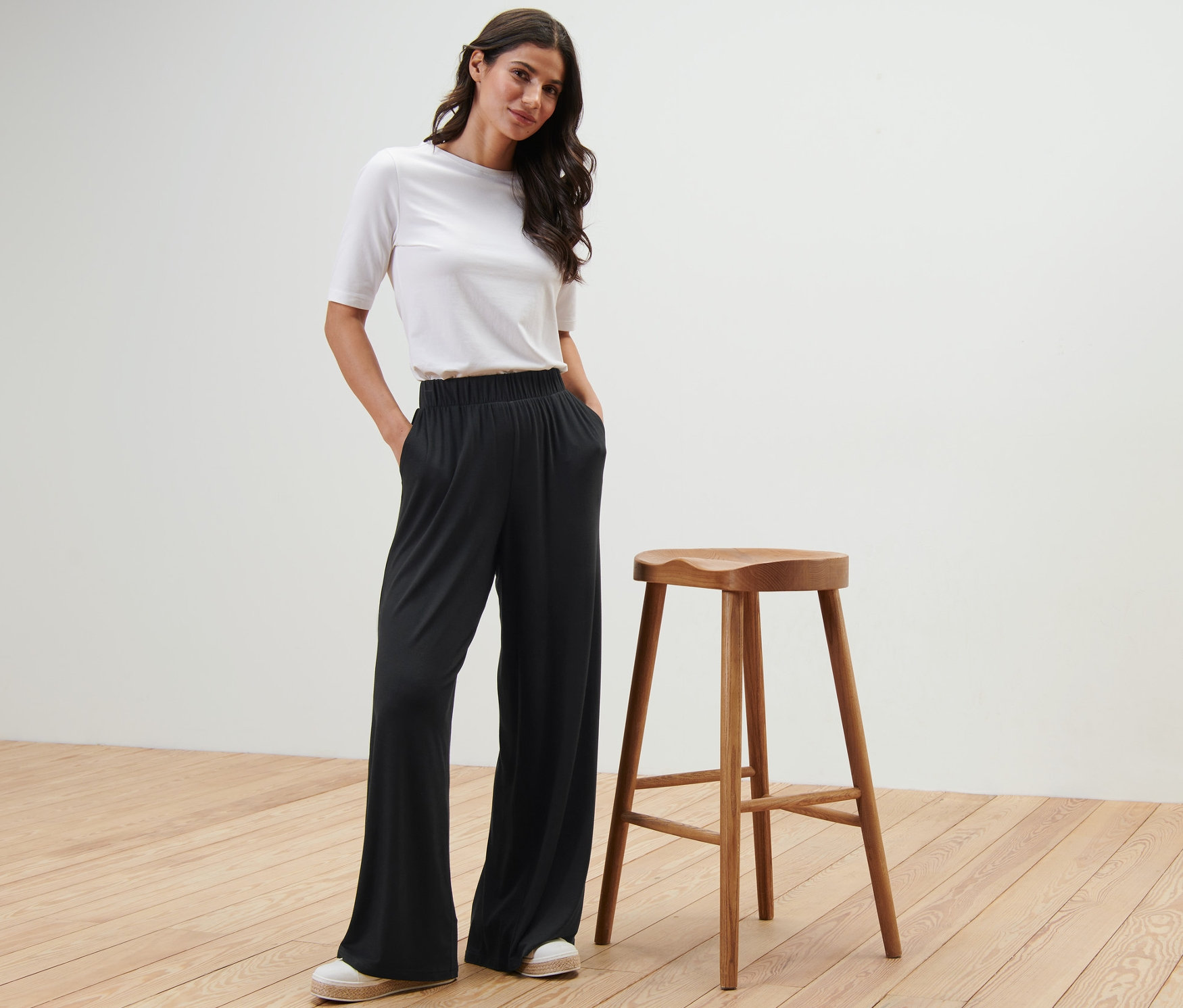 Une femme pose en pantalon palazzo noir à côté d'un tabouret en bois.