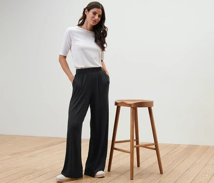 Une femme pose en pantalon palazzo noir à côté d'un tabouret en bois.