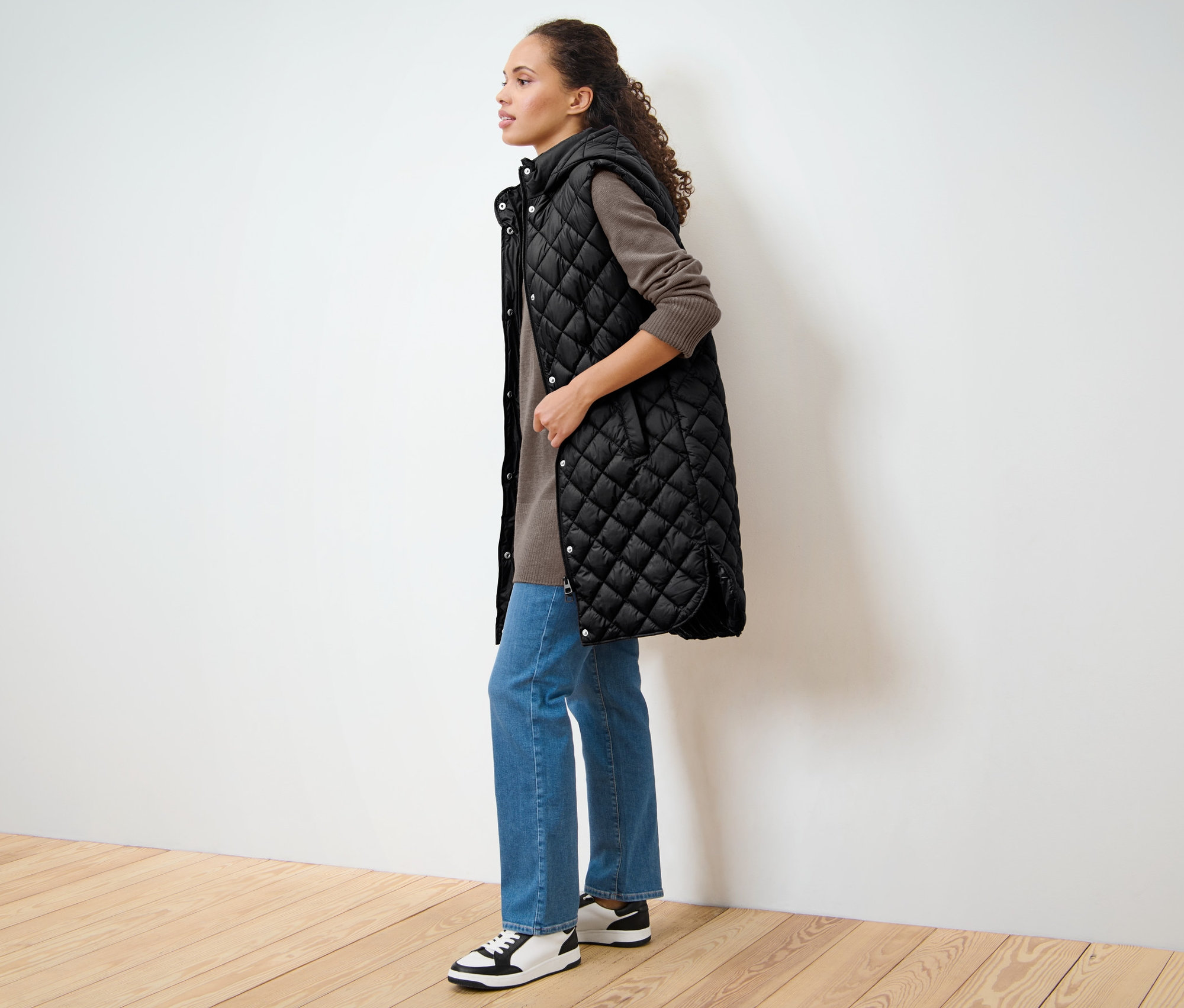 Une femme se tient de côté et porte un long gilet sans manches matelassé avec capuche.