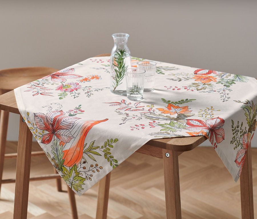 Sur une table en bois, une nappe d'environ 90 x 90 cm avec un motif floral. Une carafe d'eau et deux verres sont posés sur la nappe.