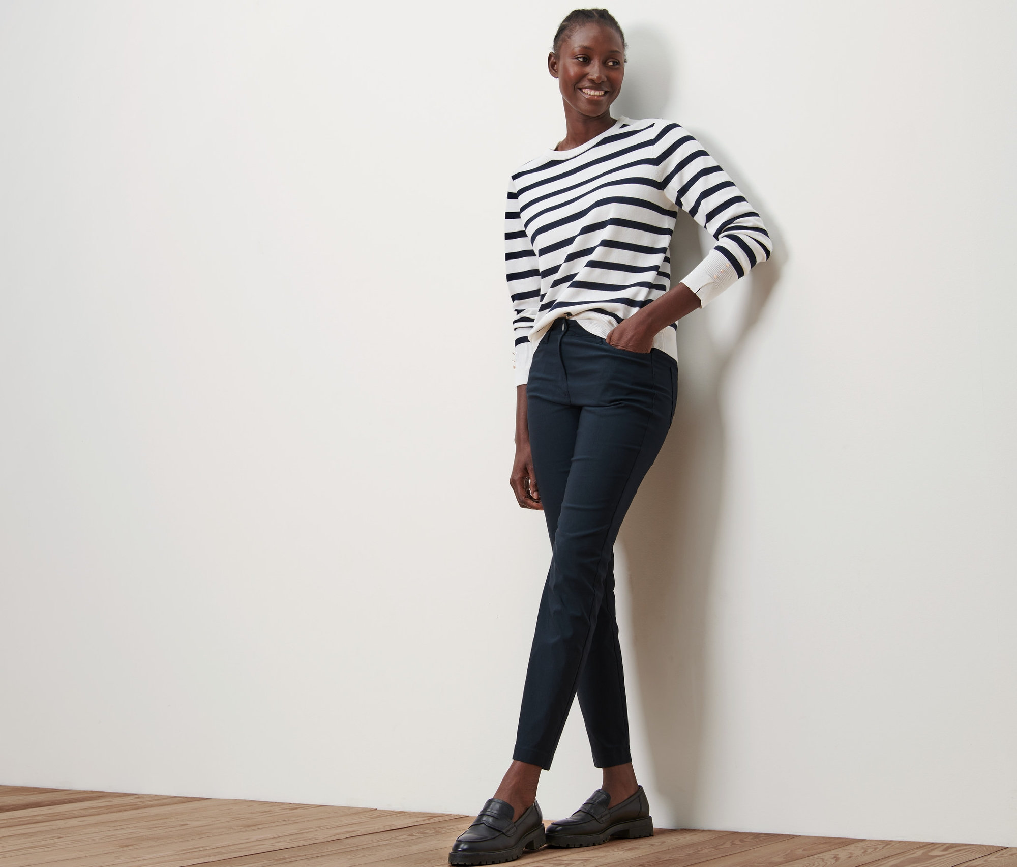 Une femme est appuyée contre un mur et porte un pantalon en bengaline bleu foncé, un haut rayé et des chaussures noires.