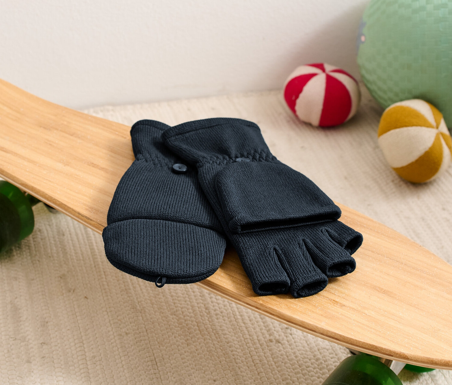 Des moufles à clapet en polaire tricotée pour enfant sont posées sur un skateboard à côté de balles.