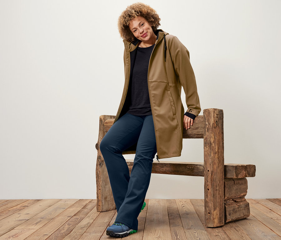 Une femme est assise sur un banc en bois et porte un manteau en softshell couleur nougat, un t-shirt de sport à manches longues noir et un pantalon de jazz thermique.