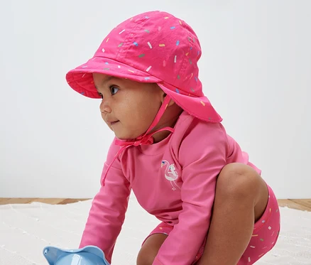 Un enfant joue avec un jouet et porte un bonnet d’été rose avec un indice de protection UV 80.