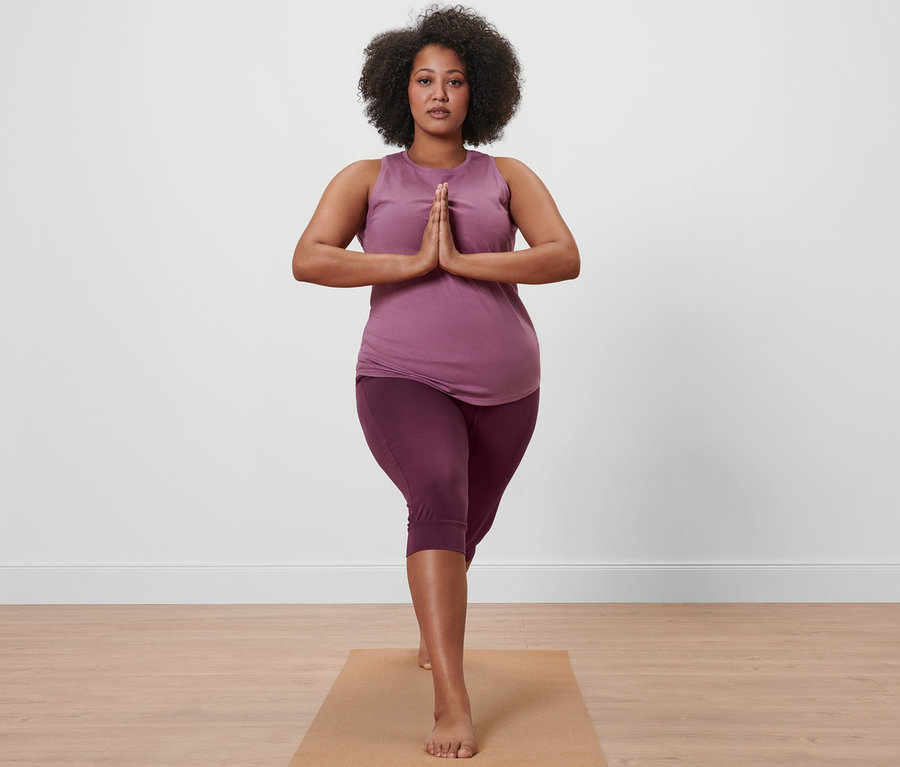 Femme en corsaire de yoga mauve et haut de sport mauve debout sur un tapis de yoga.